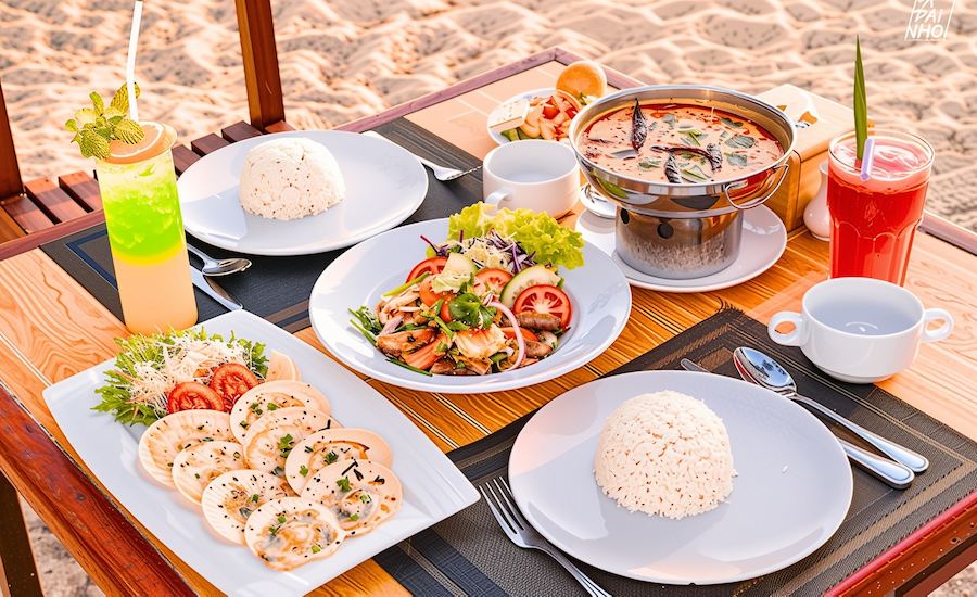 Tavolo sulla sabbia con piatti tipici thailandesi, frutti di mare e bevande colorate